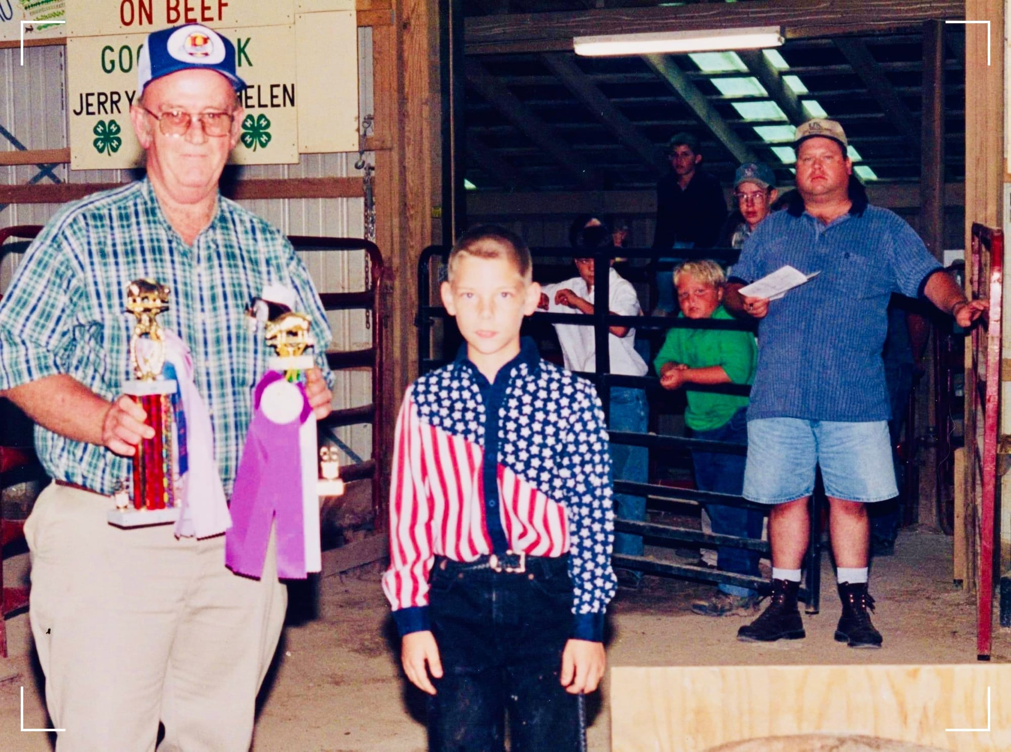 A young (boy) wearing a red, white, and blue/US-flag style shirt and black jeans, posing with an older gentleman presenting KP with two trophies during the 4-H swine show in 1999. Several adults and others look on from afar.